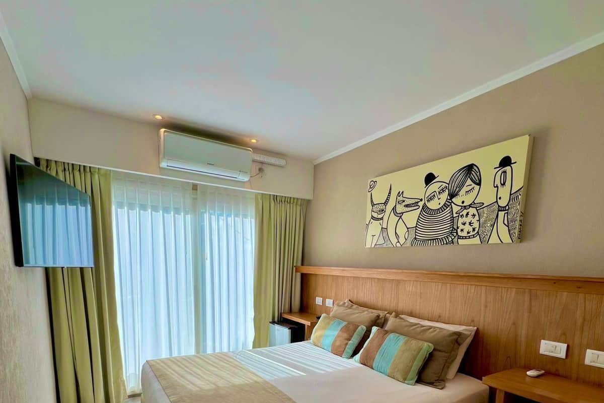 Habitación de hotel con una cama grande, un televisor de pantalla plana en la pared, aire acondicionado en el techo y un cuadro sobre la cama.