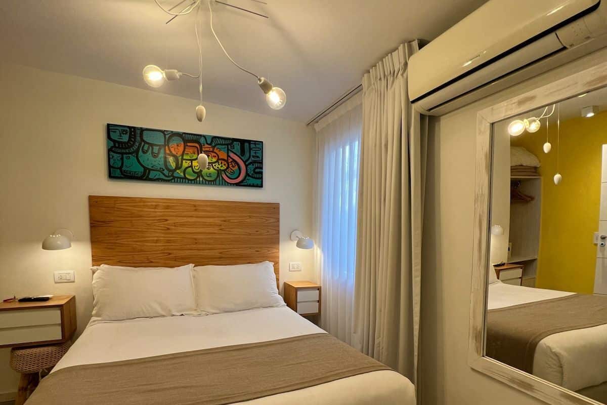 Habitación de hotel con una cama grande, lámparas de noche, una lámpara en el techo, un espejo y un cuadro abstracto colorido en la pared.