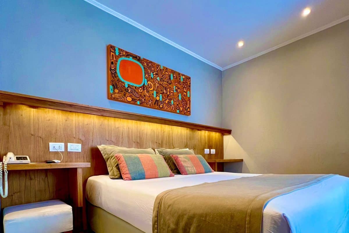 Habitación de hotel con una cama grande, cabecero de madera, mesita de noche con lámpara y una obra de arte en la pared.
