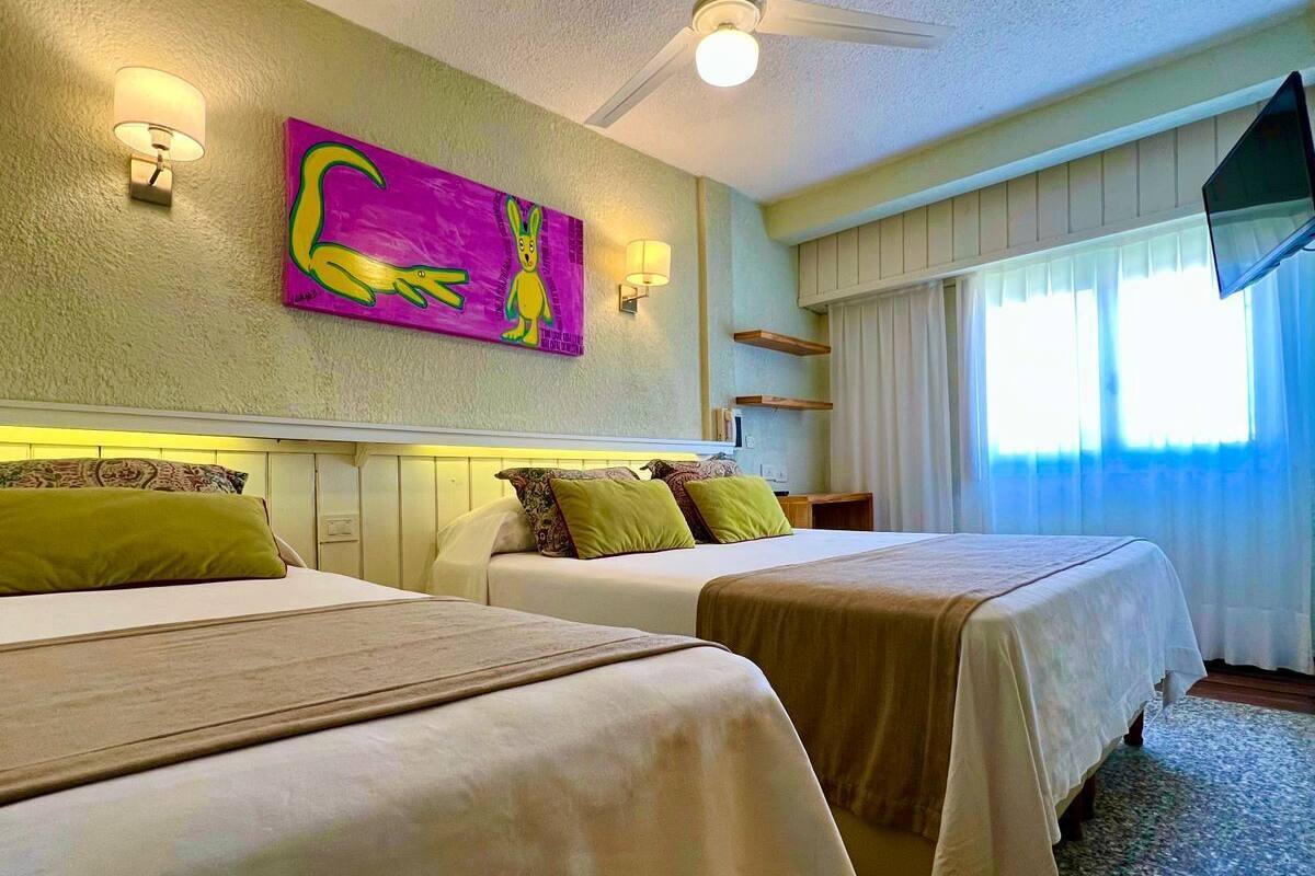 Habitación de hotel con dos camas, ventilador de techo, televisor y un cuadro en la pared.