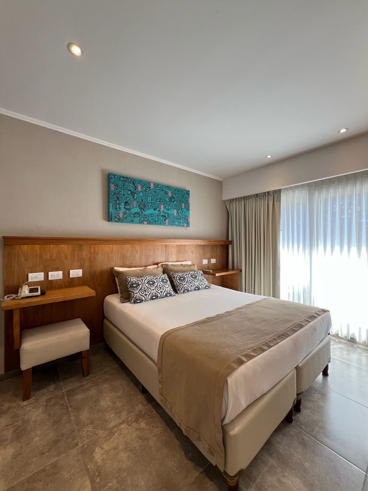 Egy szállodai szoba, amelyben egy nagy ágy, egy fafejű ágykeret, egy éjjeli asztal lámpával, egy pad és egy falra szerelt műalkotás található.