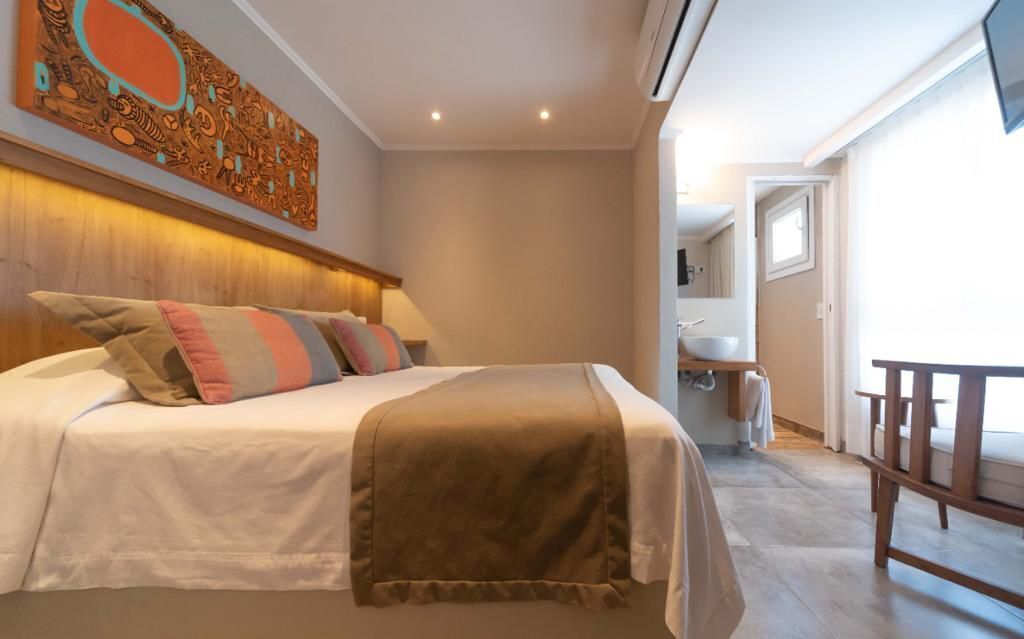 Ein Hotelzimmer mit einem großen Bett, einem hölzernen Kopfteil, einem Bild an der Wand, einem kleinen Balkon und einem Badezimmer mit Waschbecken und Spiegel.