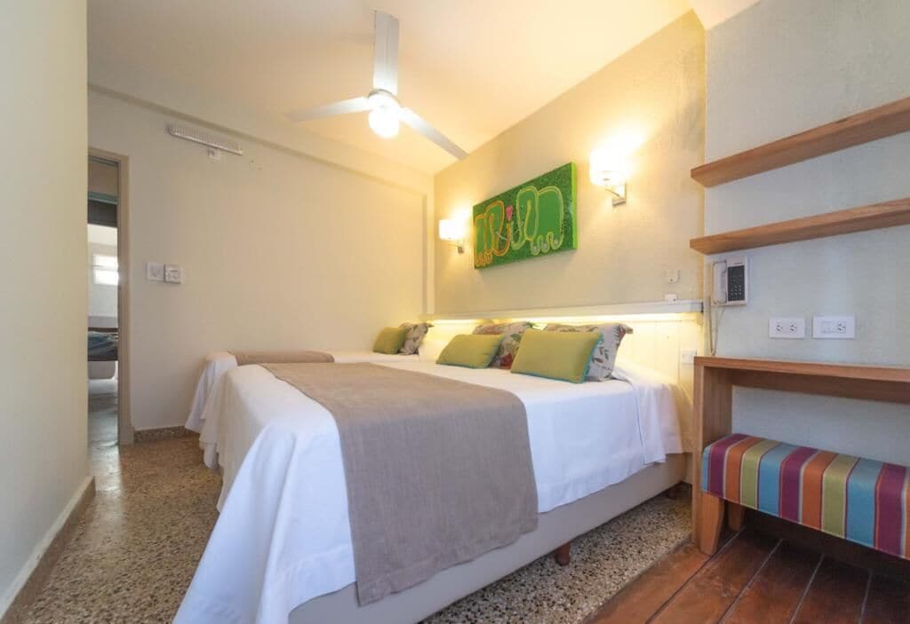 Un dormitorio con dos camas, ventilador de techo, luces fijadas a la pared, una obra de arte colorida y un banco.