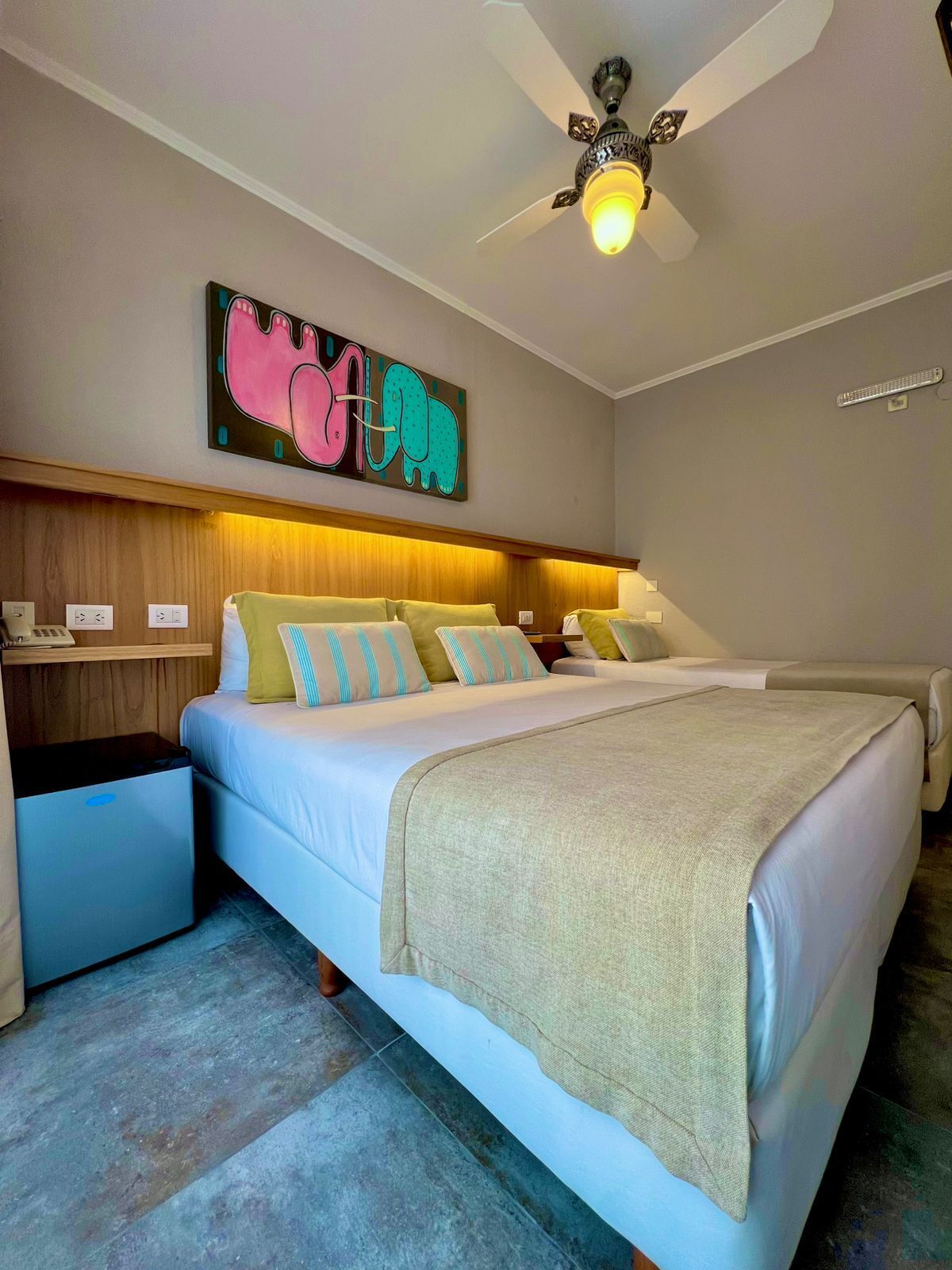 Une chambre d’hôtel équipée d’un lit, de tables de chevet, d’un ventilateur de plafond et d’une œuvre d’art accrochée au mur.