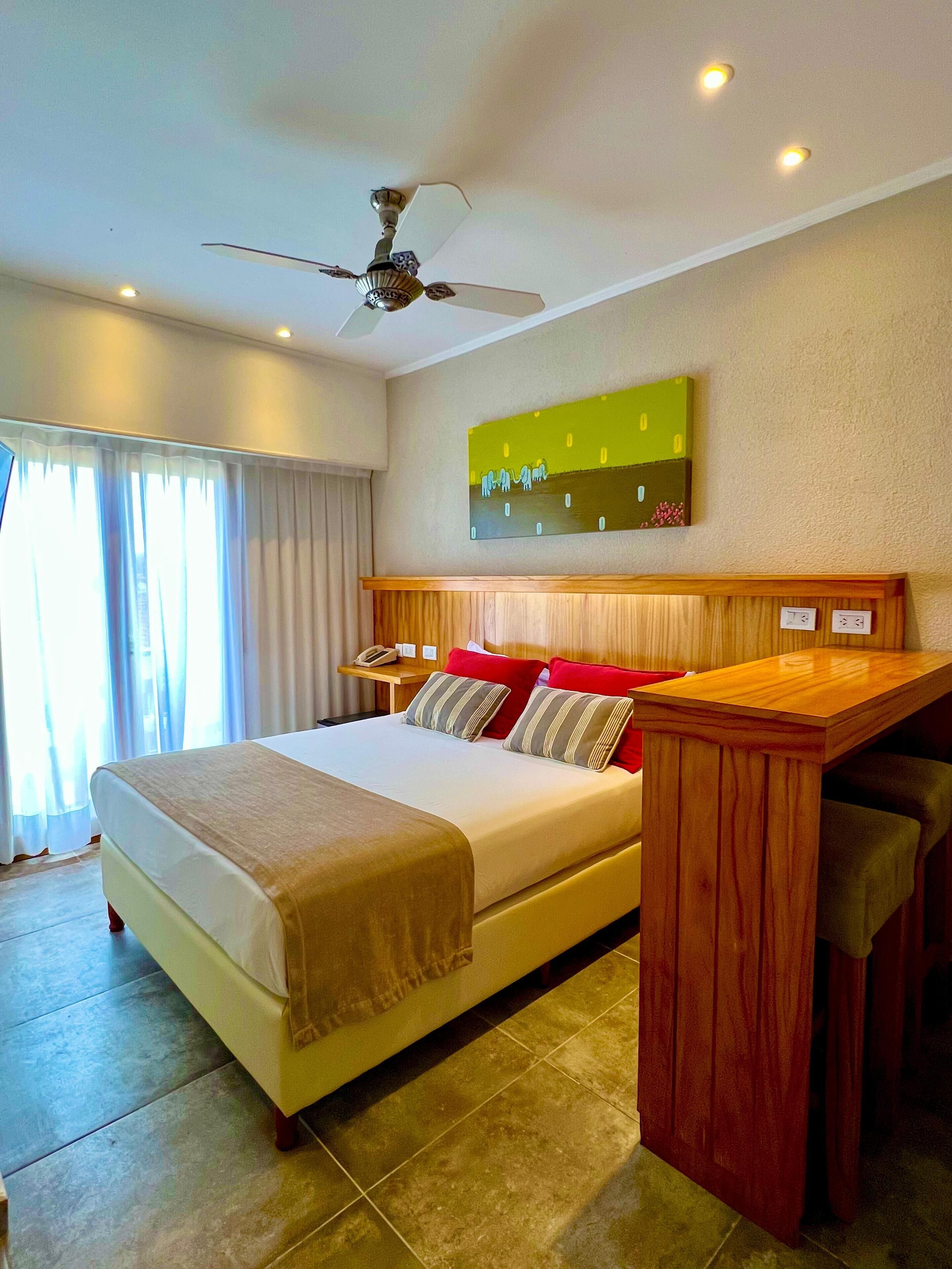 Une chambre d’hôtel avec un grand lit, une table de chevet en bois, un ventilateur de plafond et un tableau au mur.