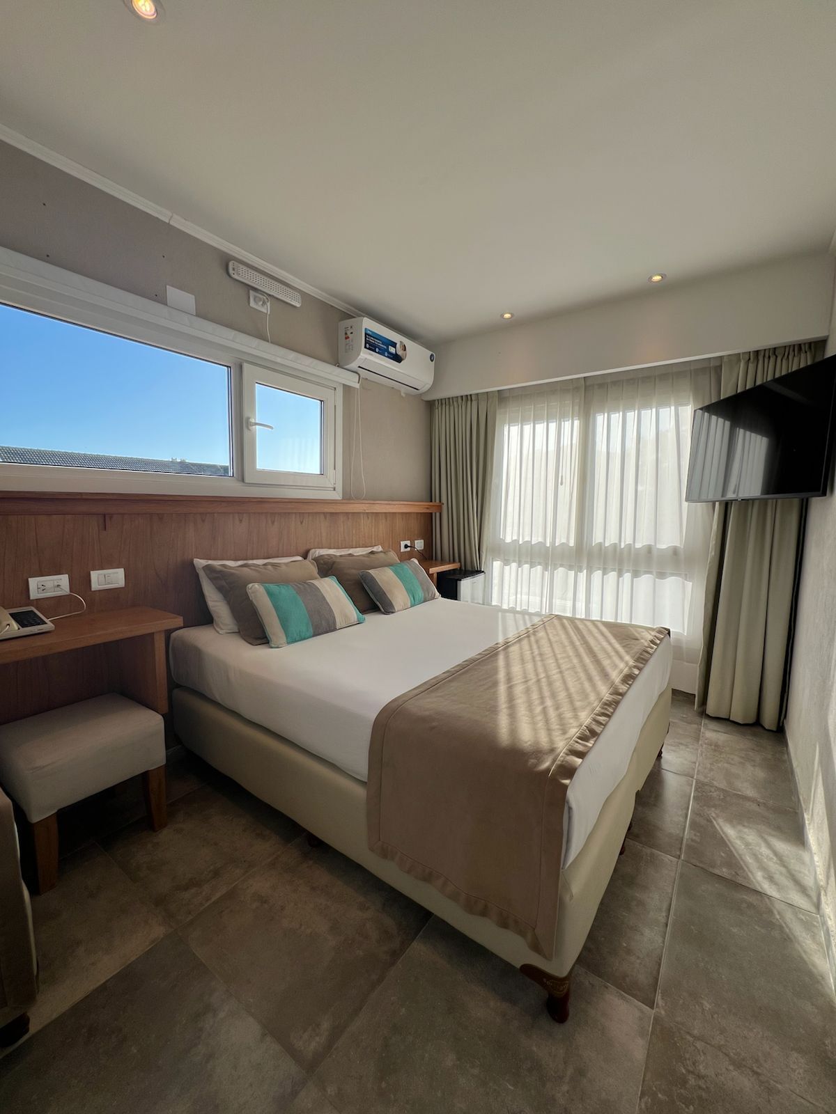 A hotel room with a bed, bedside table, chair, TV, air conditioning unit, and a window with curtains.