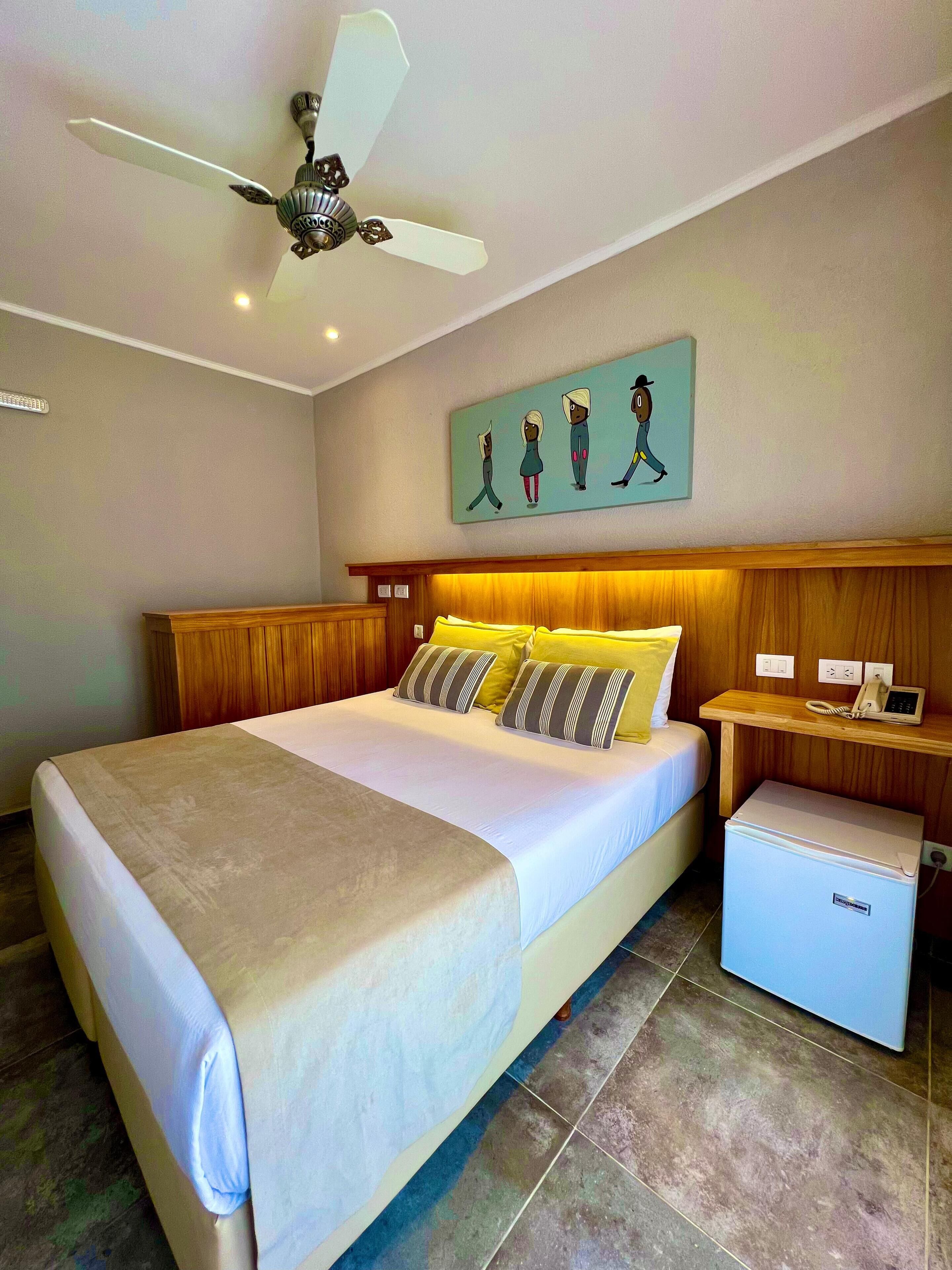 A hotel room with a bed, a ceiling fan, a small refrigerator, a bedside table with a phone, and a wall painting.