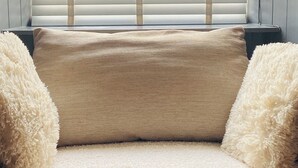 A window with horizontal blinds, a beige sofa with fluffy pillows, and a view of greenery outside. - Cosy Cottage in South Yorkshire Village. 
 (Darfield)