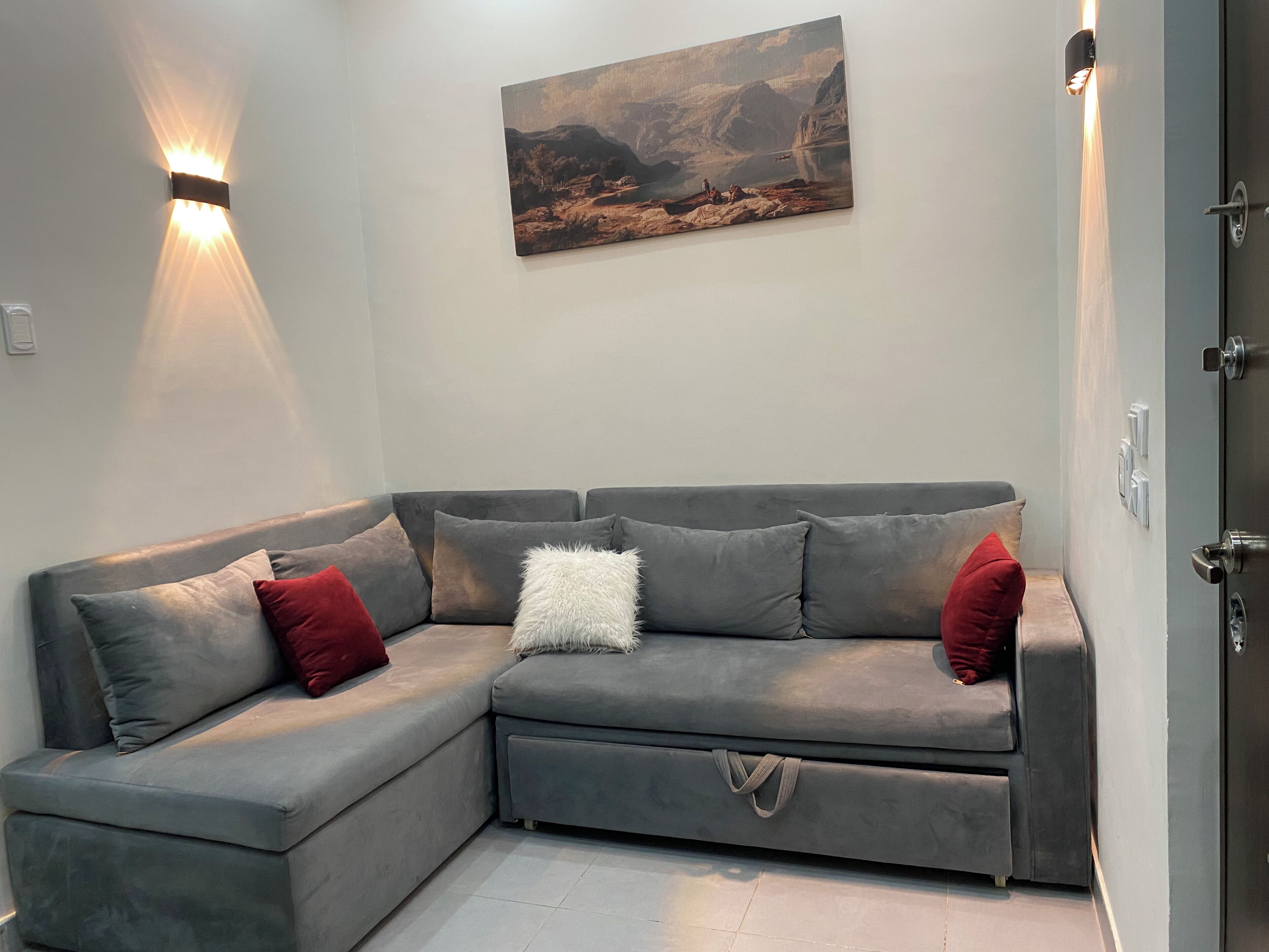 A grey sectional sofa with red and white cushions, a painting above it, and a wall-mounted light fixture.