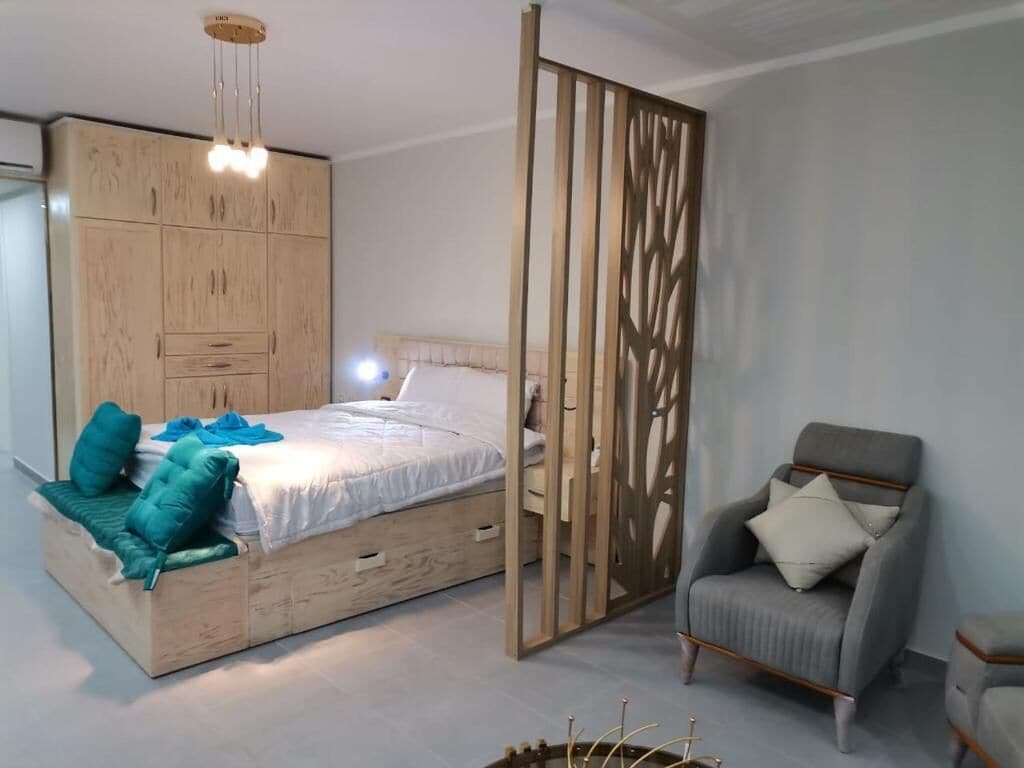 Ein Schlafzimmer mit einem Bett, einem Holzschrank, einem grauen Sessel und einem dekorativen Raumteiler.