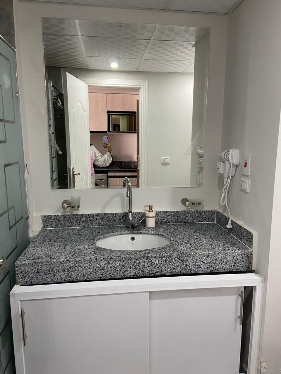 A bathroom with a granite countertop sink, a large mirror, and a hairdryer on the wall.