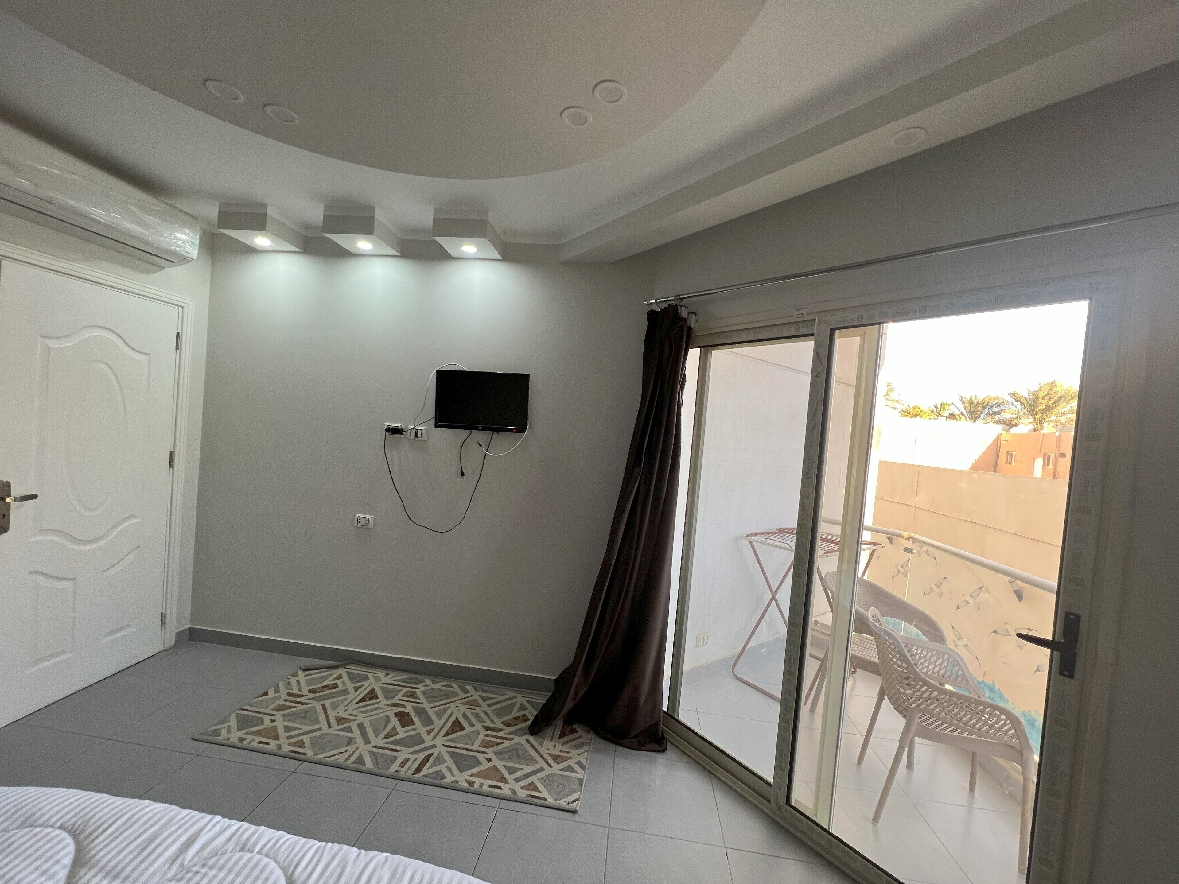 Habitación de hotel con un gran ventanal corredera a un balcón, un televisor de pantalla plana en la pared y una alfombra estampada en el suelo.