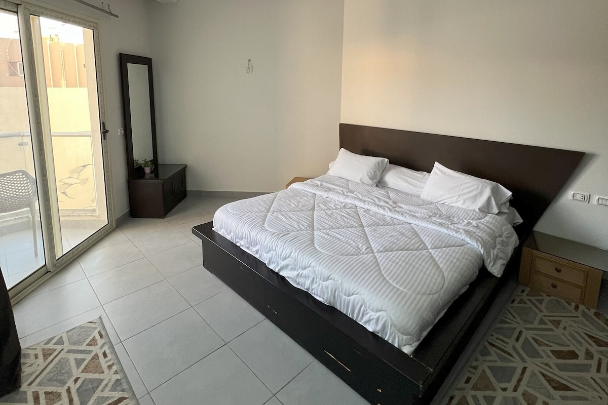 A modern bedroom with a large bed, a ceiling light fixture, and a sliding glass door leading to an outdoor area.