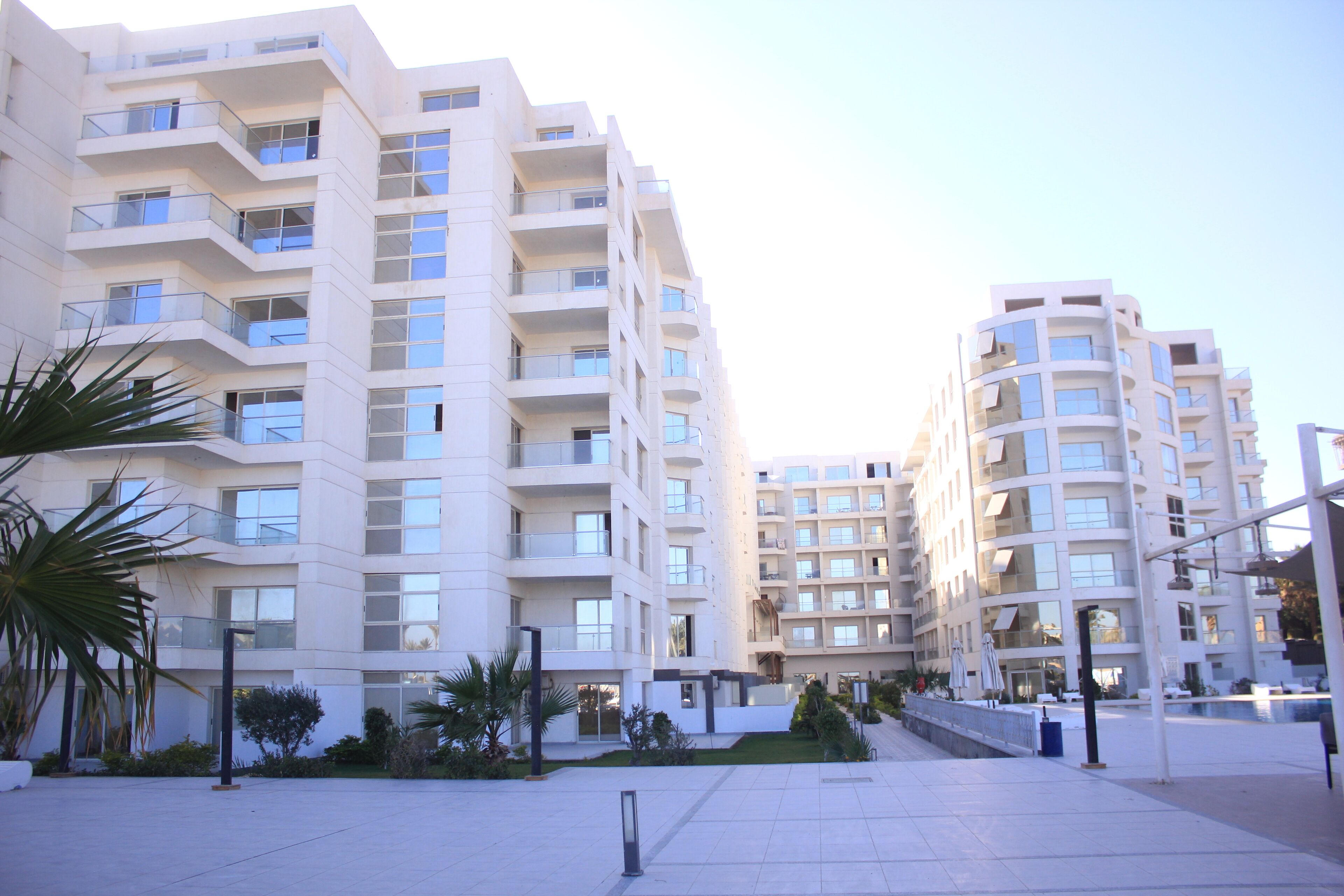 Un immeuble résidentiel moderne à plusieurs étages, doté de balcons, d’une cour aménagée et d’un espace piscine.