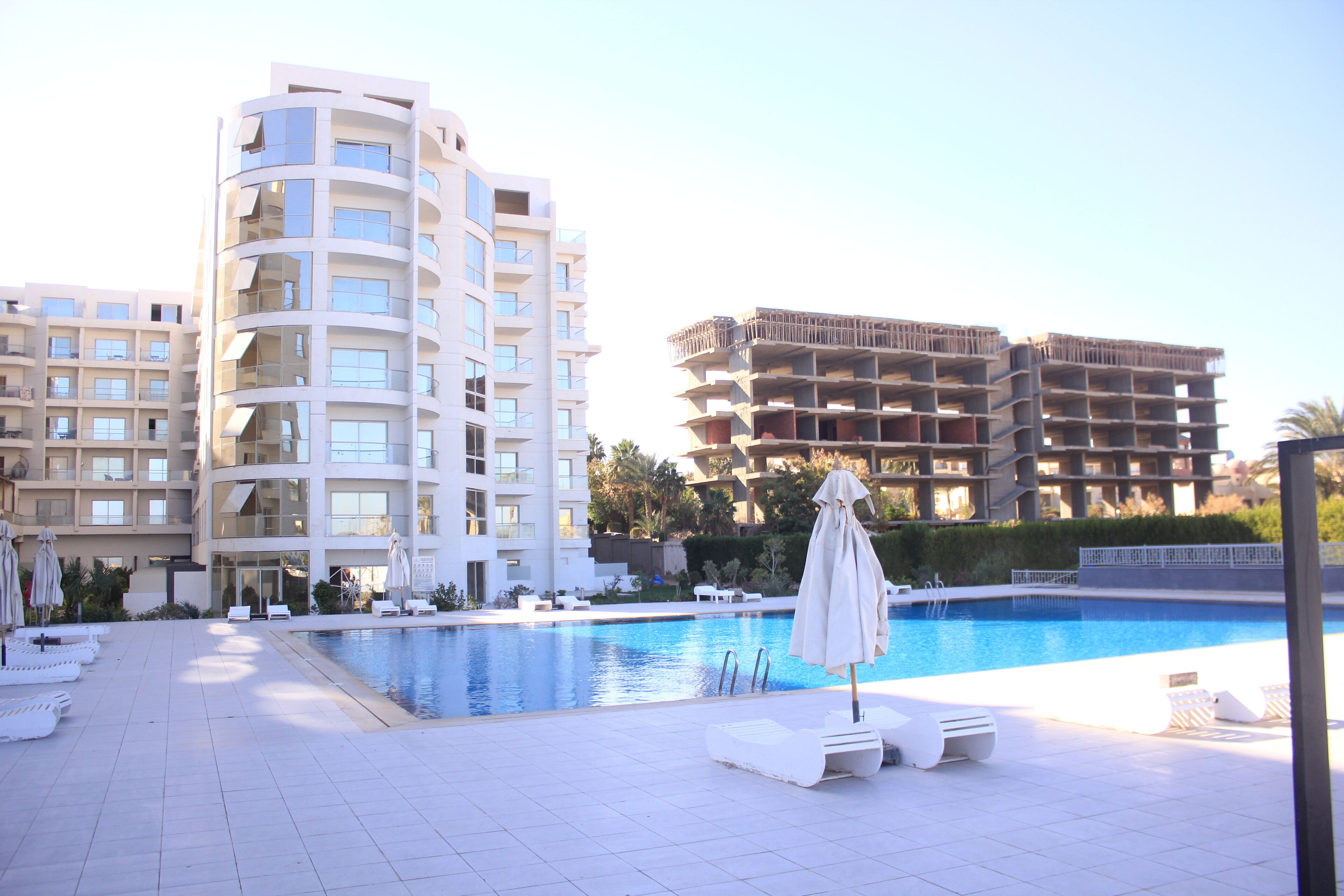 Zona de piscina con sillas de descanso y sombrillas, rodeada de edificios de varios pisos.