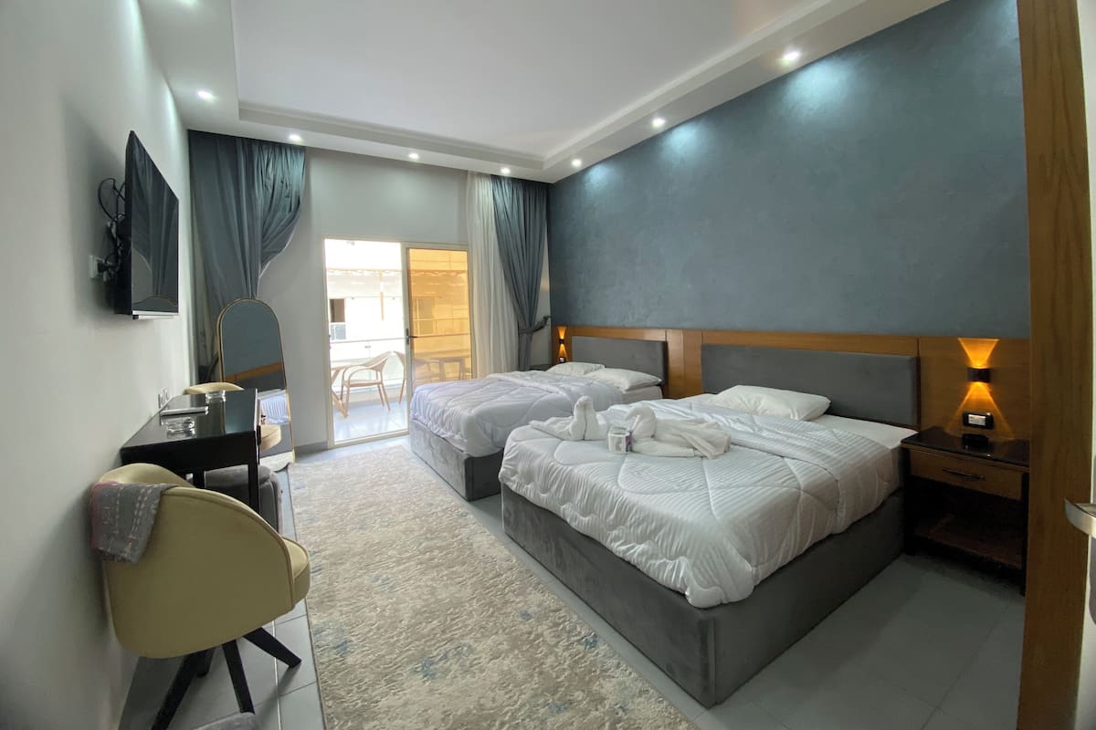 A hotel room with two beds, a desk, a chair, a TV, and a sliding door to a balcony.