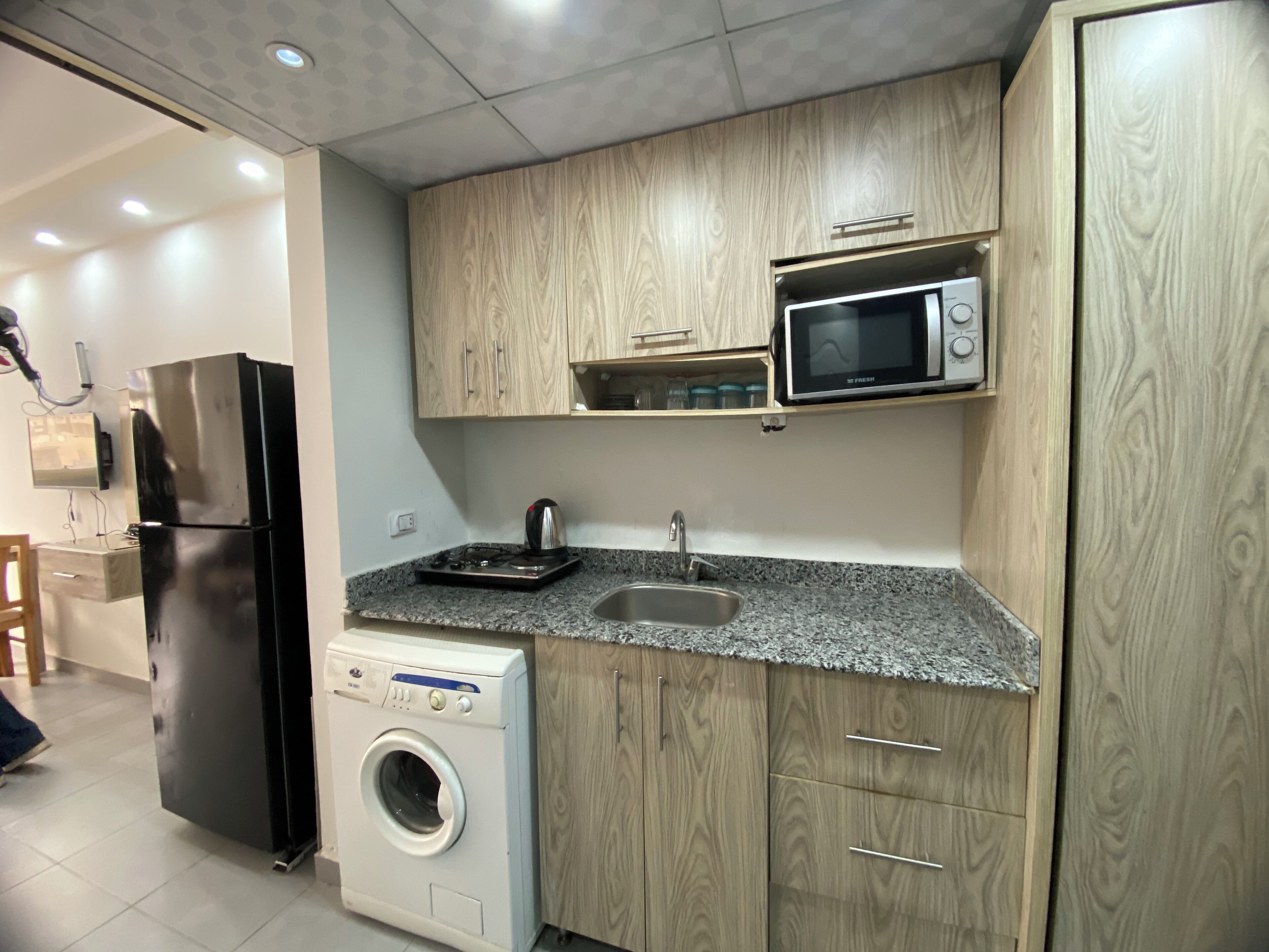 Una cucina compatta con mobili in legno, forno a microonde integrato, frigorifero in acciaio inossidabile, lavatrice e lavello.