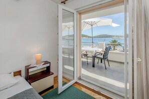 A hotel room with a bed, a nightstand, a lamp, and a sliding glass door leading to a balcony with a table, chairs, and a view of the sea. - Seafront apartments Marija with terrace and mooring (Rogoznica)