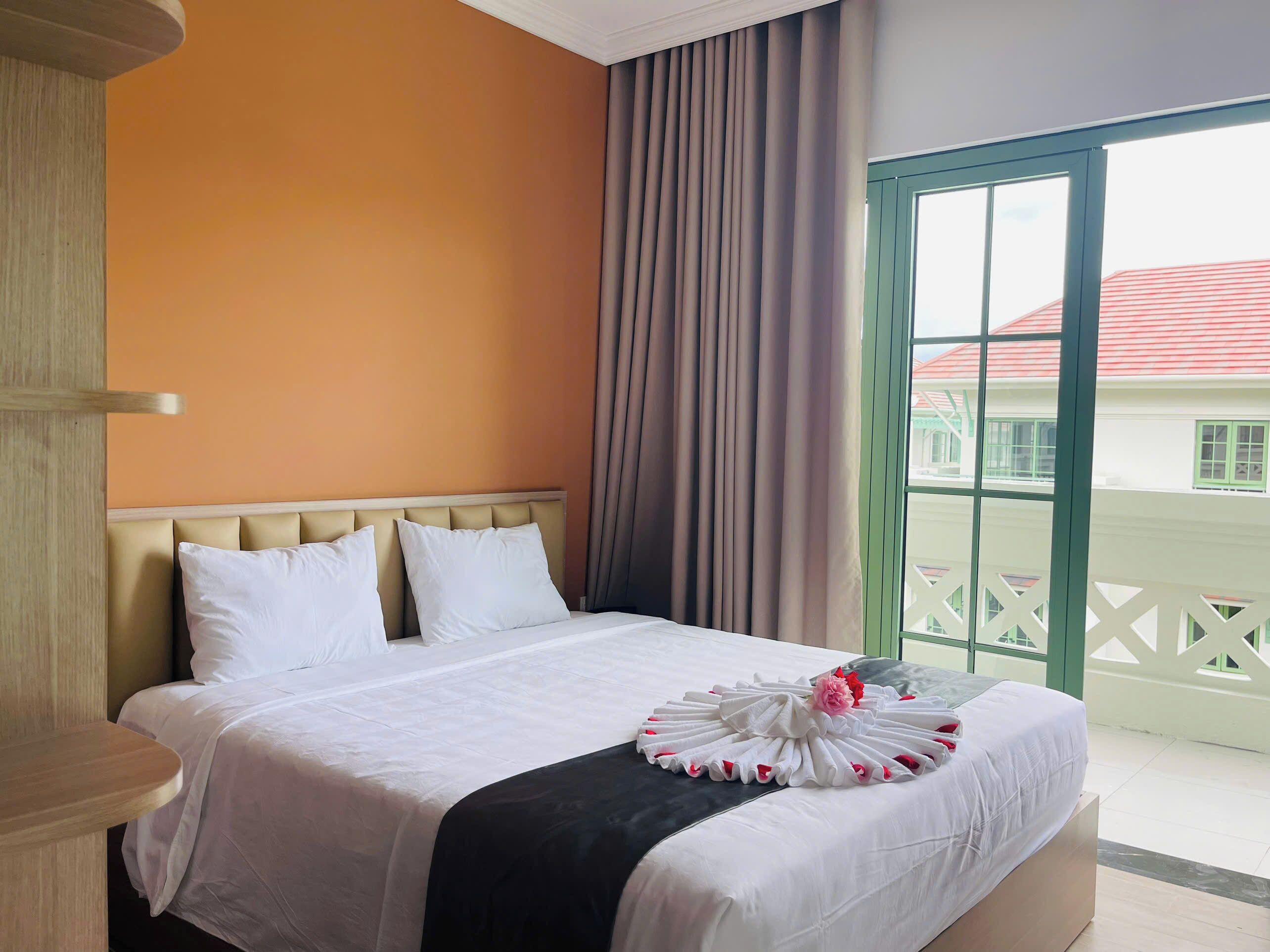 A hotel room with a bed, a wooden headboard, a window with curtains, and a balcony with a railing.