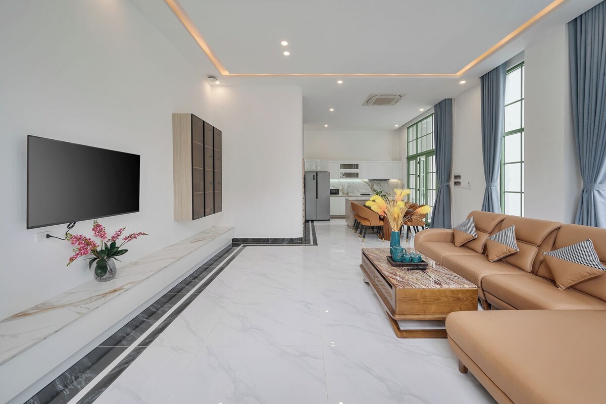 A modern living room with a flat-screen TV, a sofa, a coffee table, and a dining area in the background.