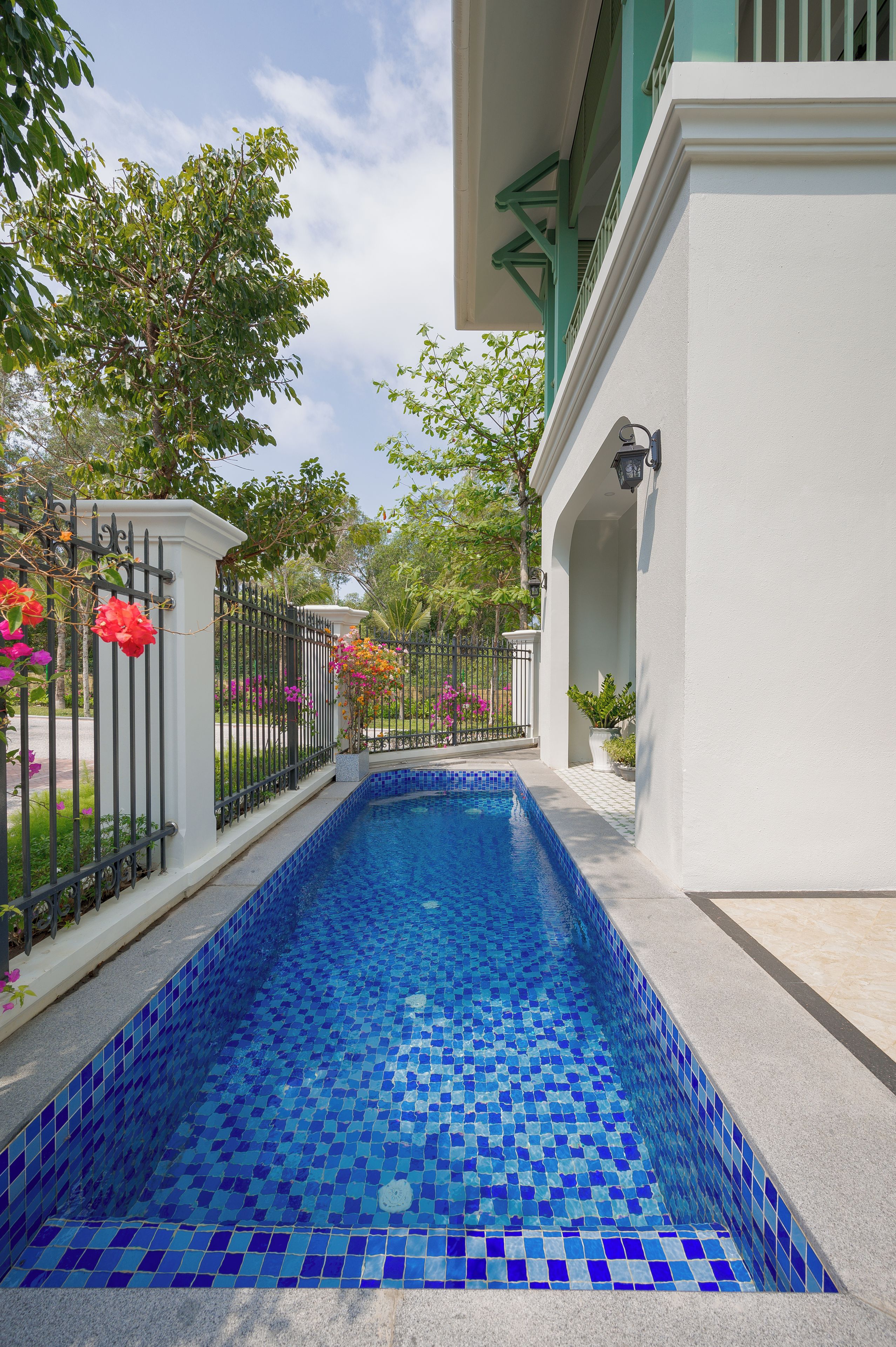 Piscina com azulejos azuis, rodeada por um edifício branco com varanda, e uma cerca de ferro preto com flores cor-de-rosa.