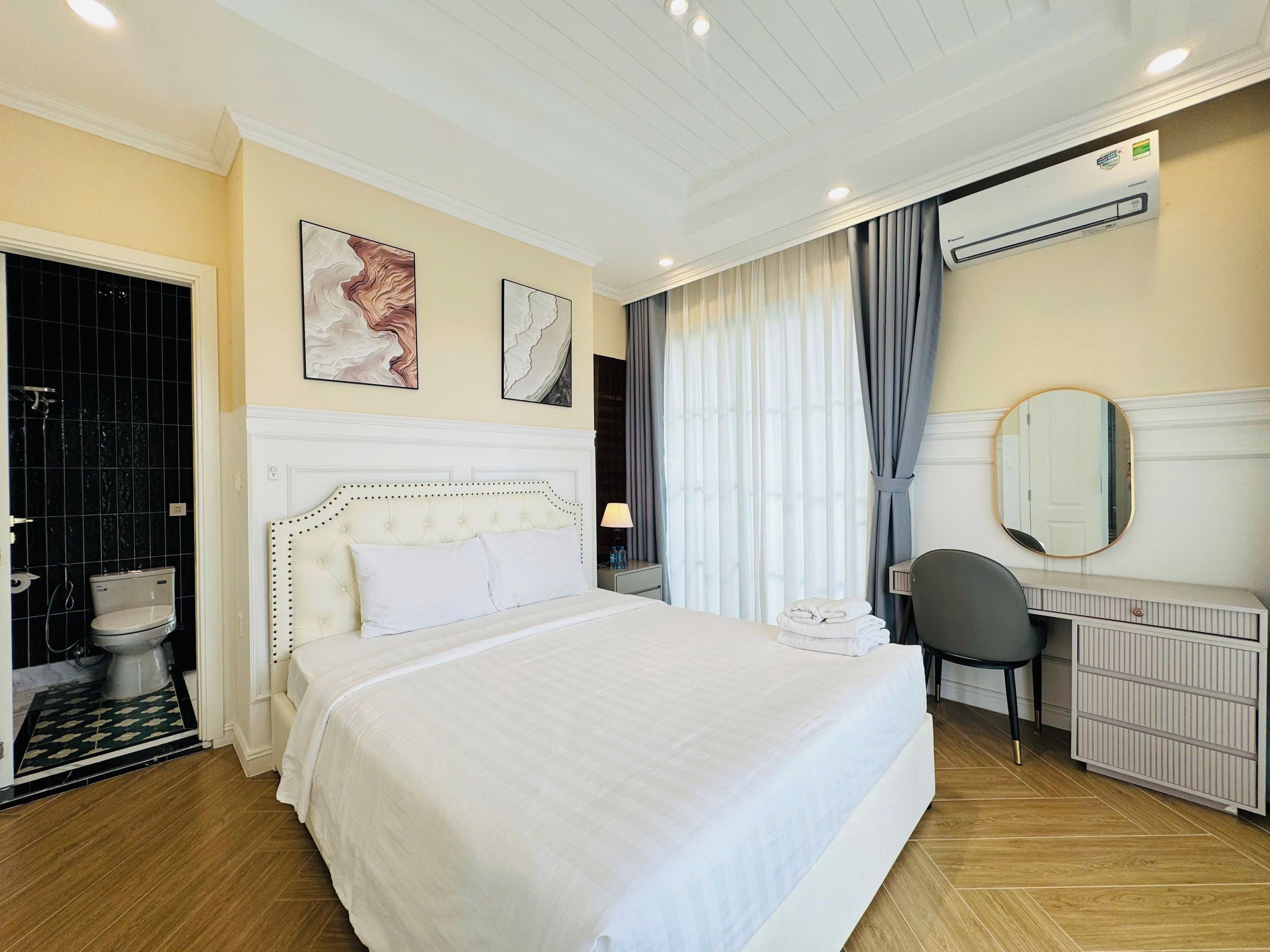 Habitació d'hotel amb un llit gran, una taula de nit, una cadira, un mirall i una finestra amb cortines.