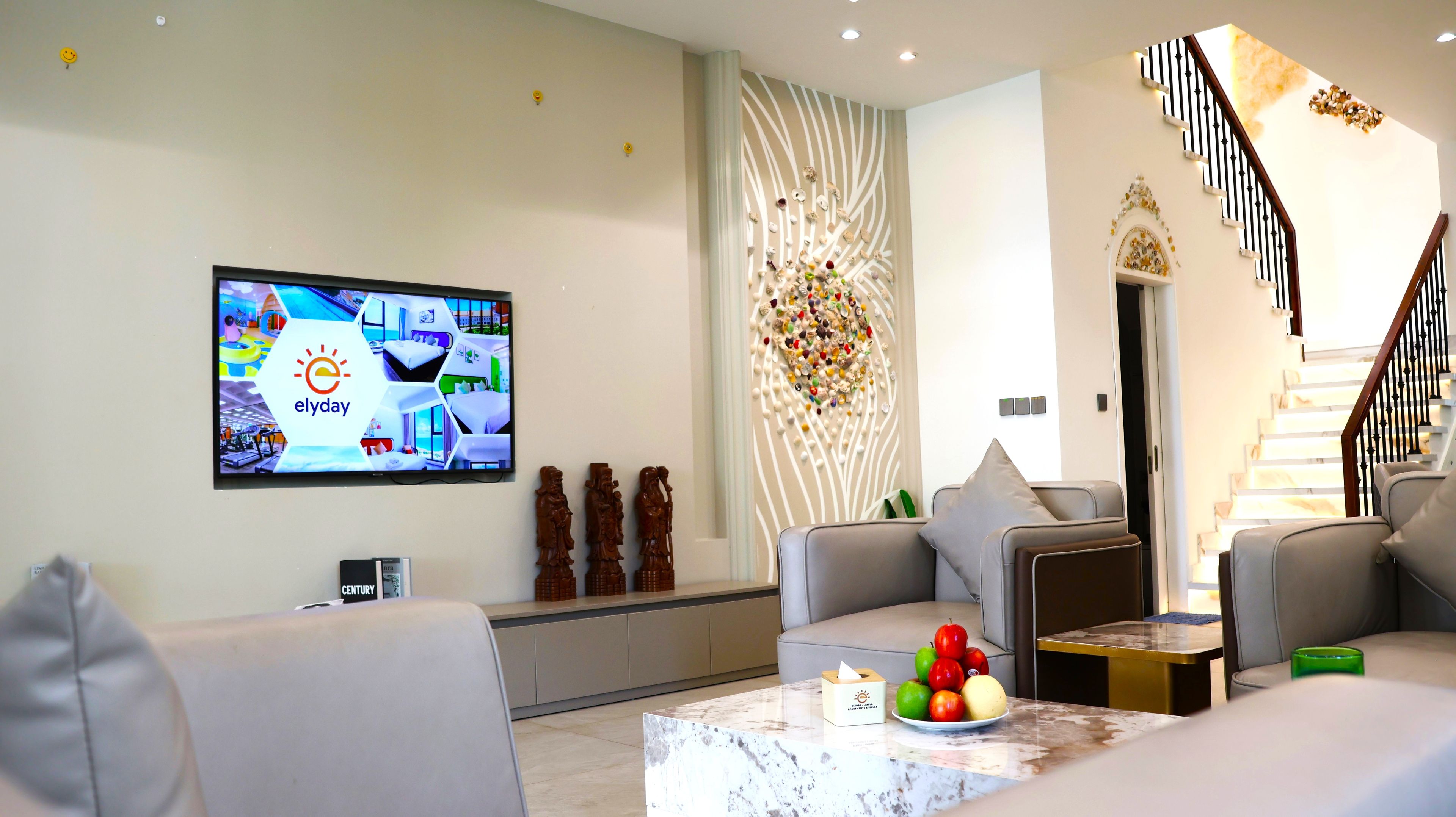 A modern living room with a flat-screen TV, a sofa, a coffee table with fruit, and a staircase.
