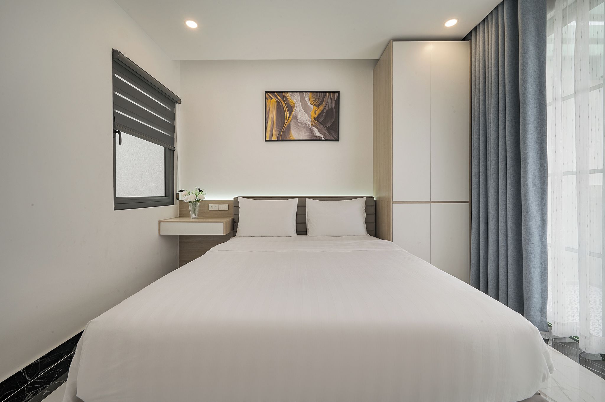 Een hotelkamer met een groot bed, een nachtkastje met een vaas, een schilderij aan de muur en een raam met gordijnen.