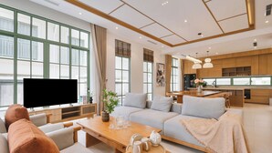 A modern living room with a large flat-screen TV, a sofa, a dining table, and a kitchen area with wooden cabinets and pendant lighting. - Elyday Villa - Bai Khem Beach, Phu Quoc (Phu Quoc)