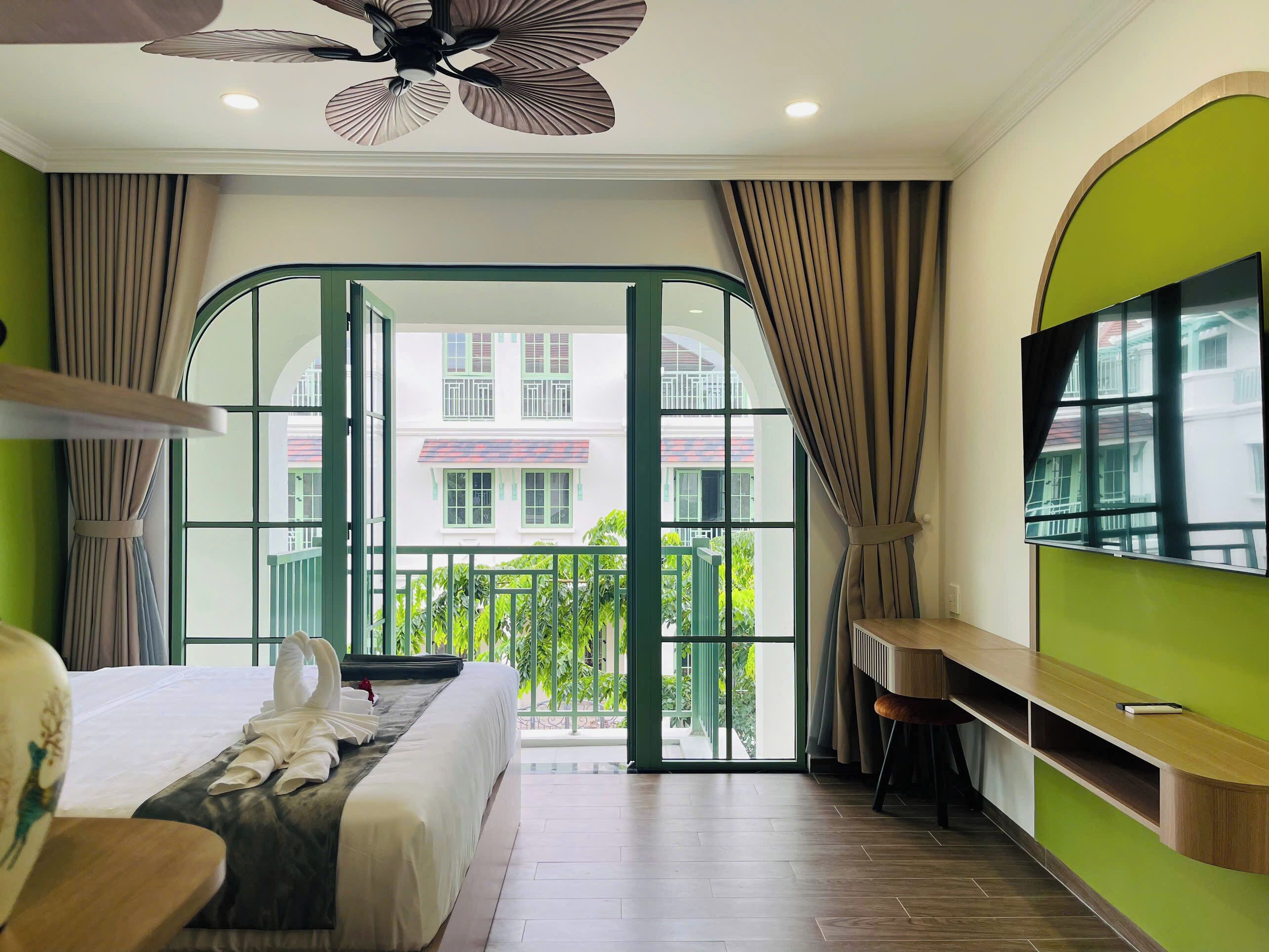 Quarto de hotel com cama, escrivaninha, TV e varanda com vista para edifícios e vegetação.