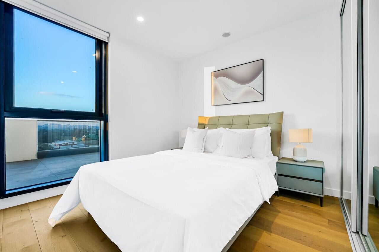 Une chambre moderne avec un grand lit, une table de chevet, une lampe et une fenêtre donnant sur la ville.