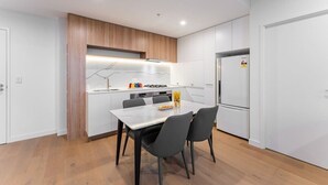 A modern kitchen with white cabinets, a wooden accent wall, a dining table with chairs, and a refrigerator. - StayAU Spacious 2BR Stunning Sunset Views (Box Hill)