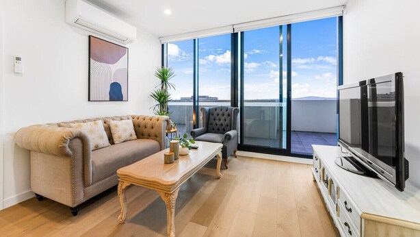 A modern living room with a sofa, armchair, coffee table, TV, and large windows offering a city view. - StayAU Spacious 2BR Stunning Sunset Views (Box Hill)