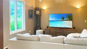 Un soggiorno con una grande TV a schermo piatto, un divano, una sveglia e una finestra con vista su spazi verdi.