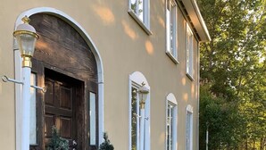 Una casa con una porta in legno scuro, finestre con cornice bianca e un giardino curato con fiori rosa e arbusti verdi.