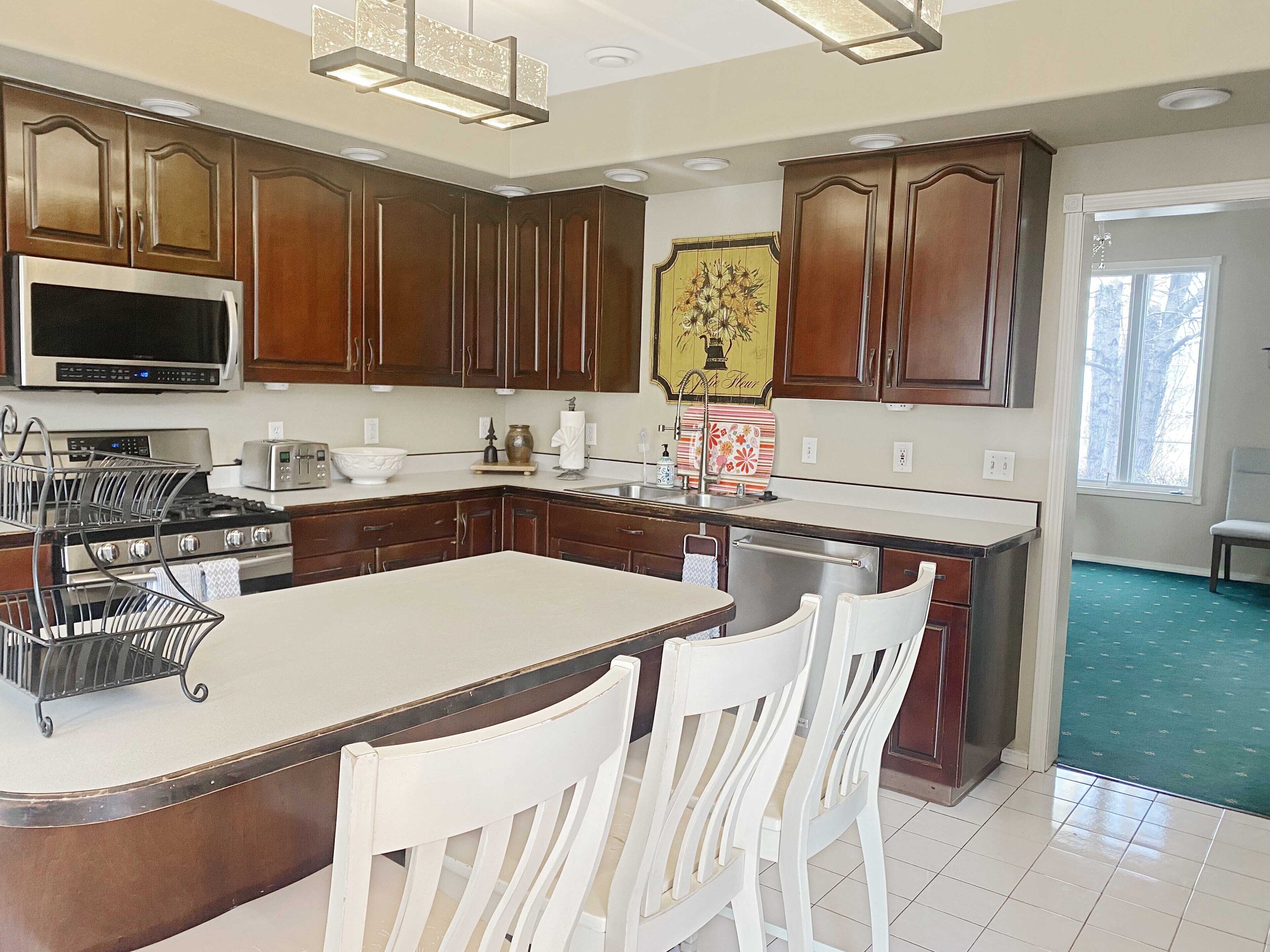 A kitchen with wooden cabinets, a stainless steel refrigerator, a microwave, a stove, and a large island with white chairs.