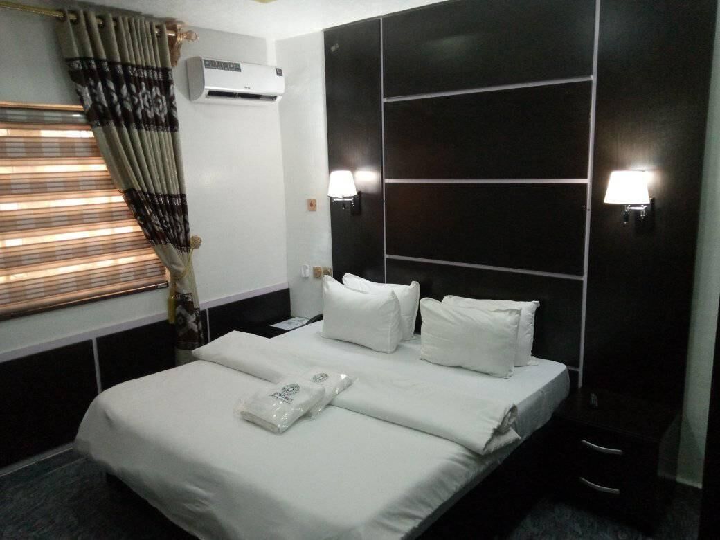 A hotel room with a large bed, white linens, a dark headboard, two wall-mounted lamps, and an air conditioning unit.
