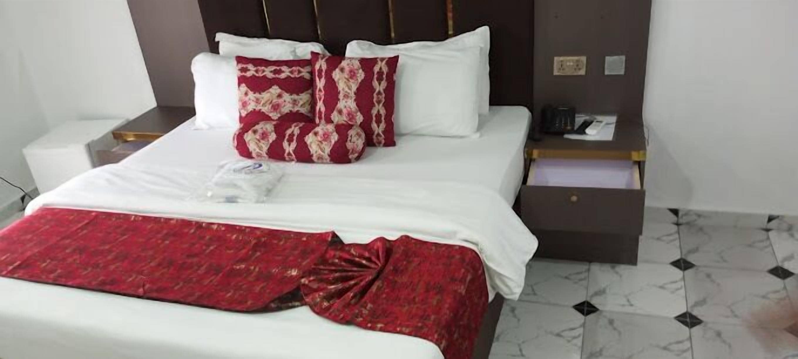 Ein Hotelzimmer mit einem Bett, weißer Bettwäsche, roten und weißen Kissen, einer roten Decke, einem Nachttisch mit Telefon und Tasse sowie einem gefliesten Boden.