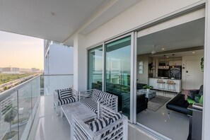 Un balcon doté d’une balustrade en verre, un coin repas avec une table et des chaises, et une cuisine visible à travers les portes vitrées.