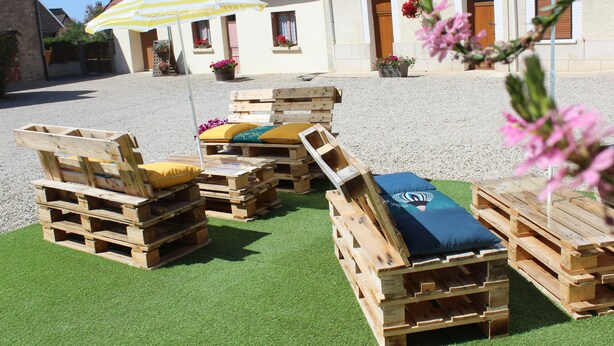 Mobiliario exterior hecho con palets de madera, con cojines, un paraguas a rayas amarillo y blanco, y un edificio de techo rojo al fondo.