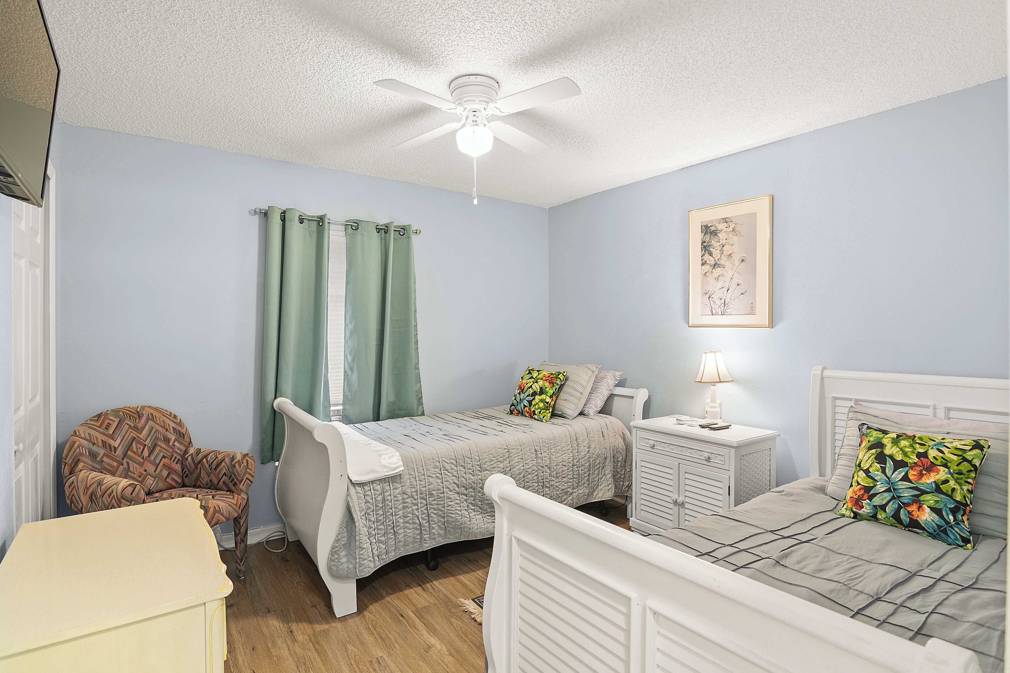 A bedroom with a bed, a chair, a nightstand, a ceiling fan, and a window with curtains.