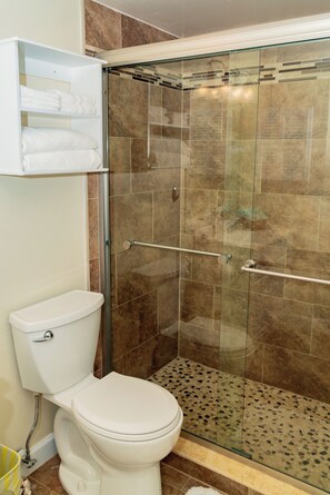A bathroom with a glass-enclosed shower, a toilet, and a shelf with folded towels.