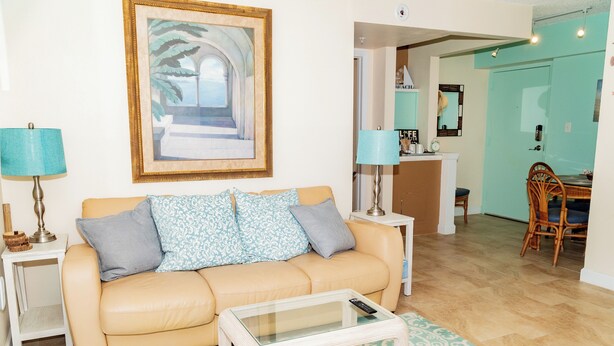 Ein Wohnzimmer mit einem beigefarbenen Sofa, gemusterten Kissen, einem Glastisch, einem türkisfarbenen Teppich und einem gerahmten Bild an der Wand.