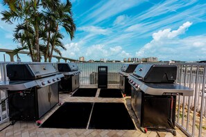A waterfront outdoor grill area with two barbecue grills, a paved walkway, and a view of the water and buildings in the background.