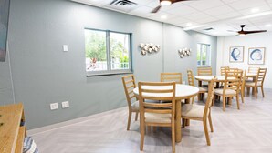 A dining area with wooden chairs and tables, a ceiling fan, and windows with views of greenery. - #803 Lovers Key Beach Club Ocean Views (Bonita Springs)