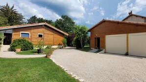 A two-story wooden house with a gravel driveway, a garage, and a small garden with potted plants. - Reflet de Bien être : Wooden loft for 3 people with enclosed courtyard. (Marsoulas)