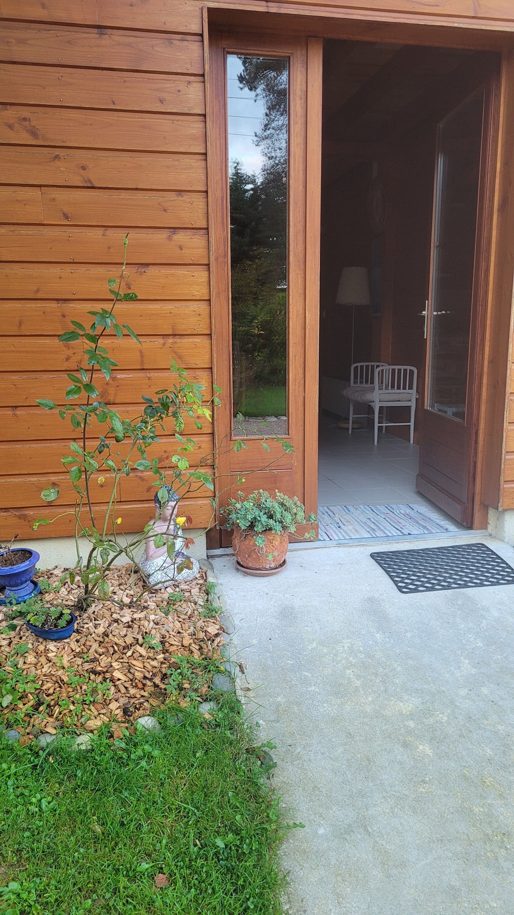 A wooden building with a glass door leading inside. There is a small garden with plants and mulch in front.