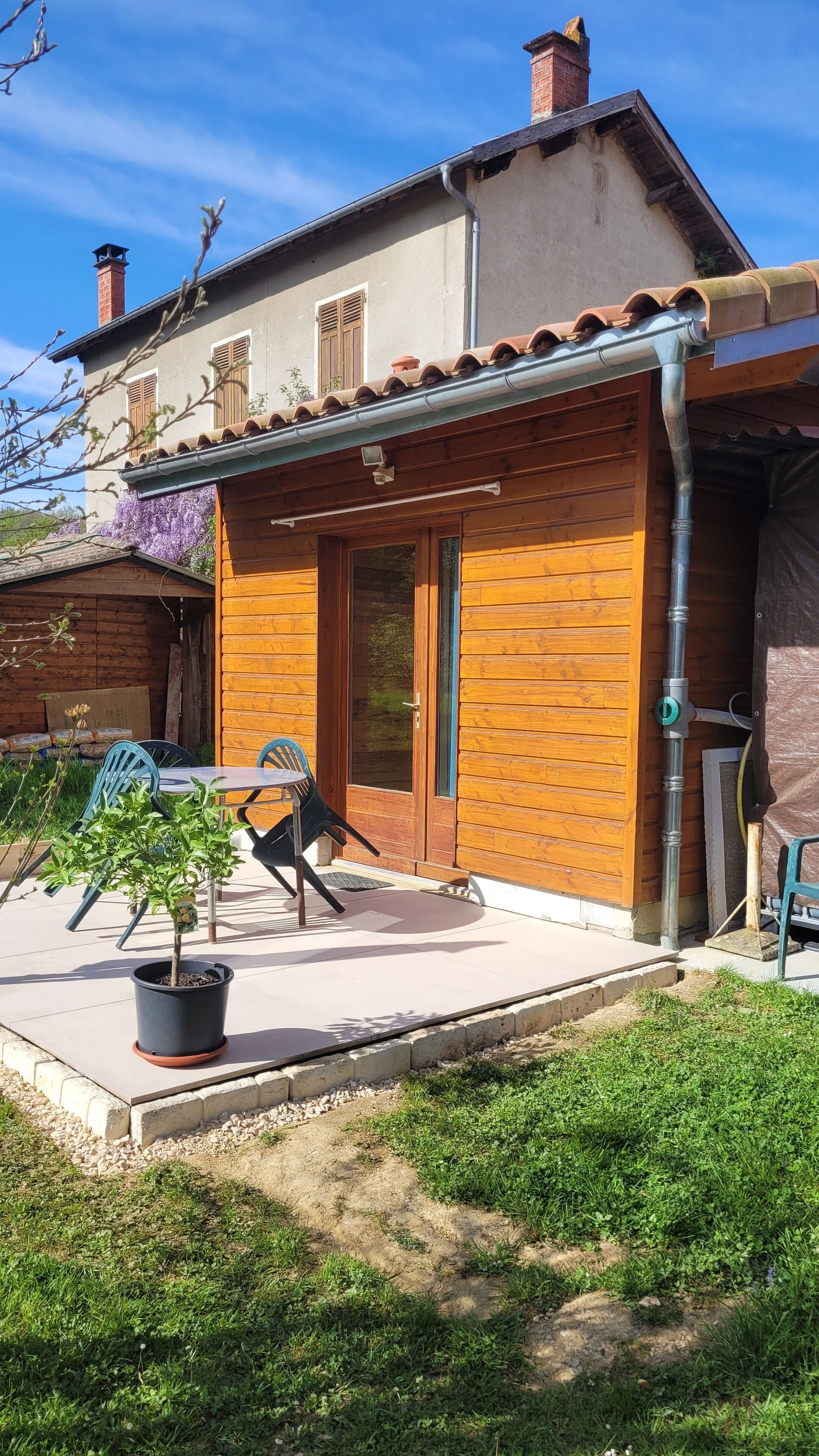 Una casa de madera con techo de tejas, una puerta principal y un pequeño patio con mesa y sillas.