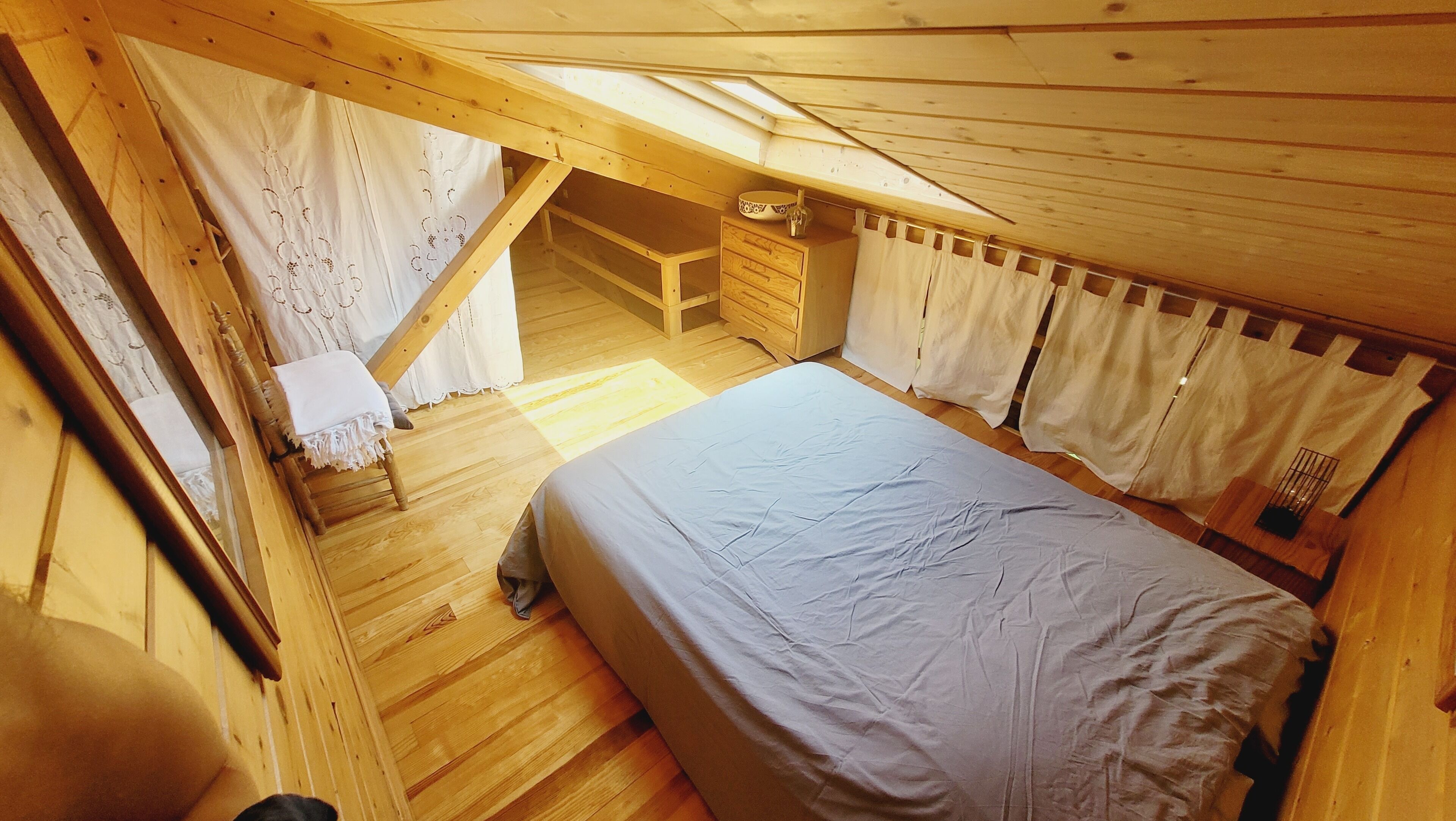 Een slaapkamer in een houten chalet met een hoogslaper, een klein houten bankje en een raam met gordijnen.