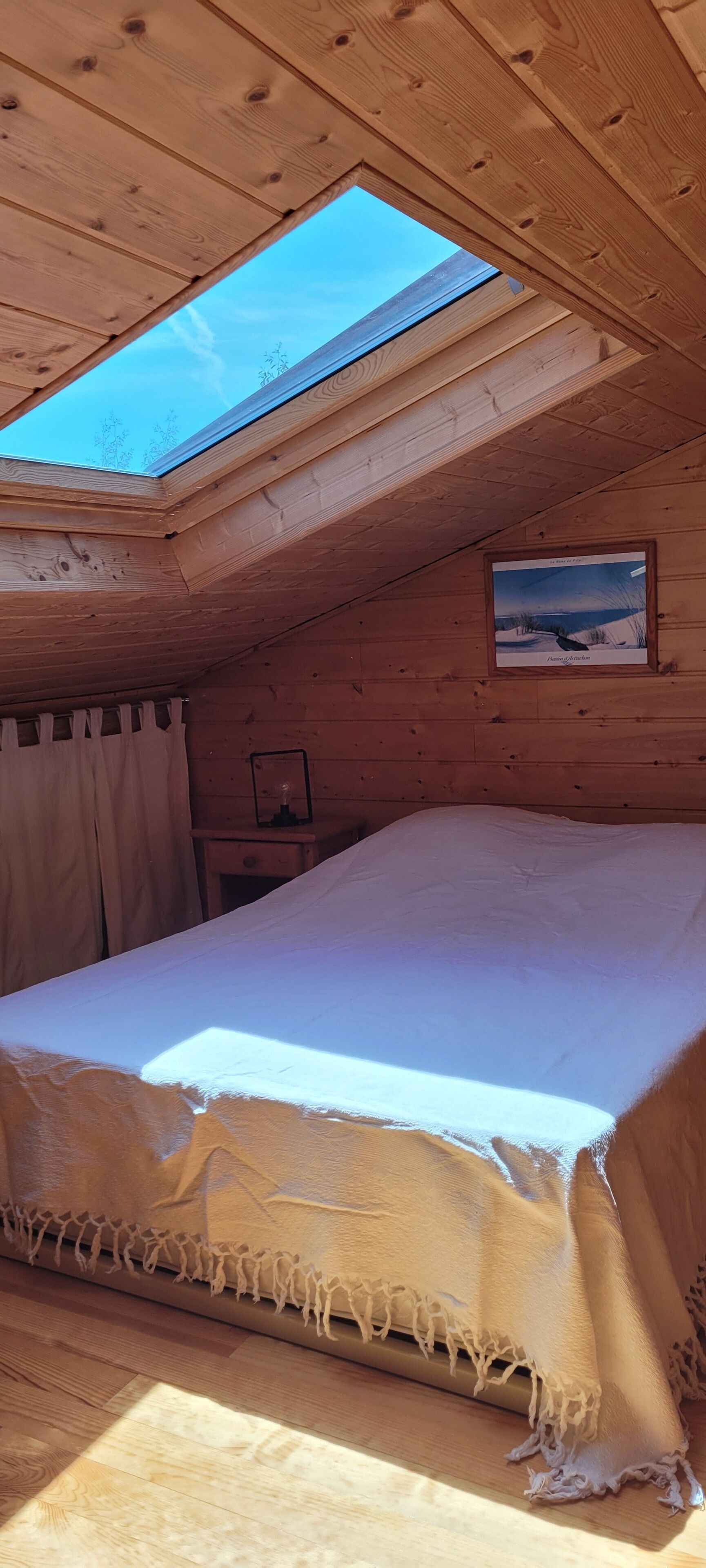 A cozy room with a skylight, a bed with a white sheet, a framed picture on the wall, and a wooden nightstand.